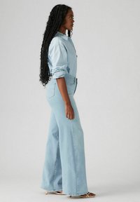 Lichtblauwe denim shirt en wijde jeans. Losse pasvorm, opgerolde mouwen en gestructureerde stof. Kleuren passen bij elkaar en worden aangevuld door lichte sandalen.