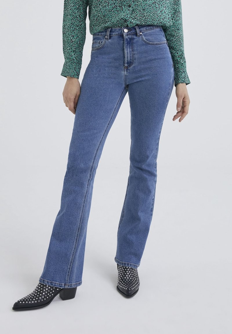 Femme portant un jean bleu taille haute, une chemise verte à manches longues à motifs et des bottines noires à talons cloutées, debout devant un fond uni.