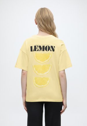Personne aux longs cheveux blonds porte un t-shirt jaune avec l'inscription « FRESH VIBRATIONS LEMON » et trois imprimés de tranches de citron dans le dos.