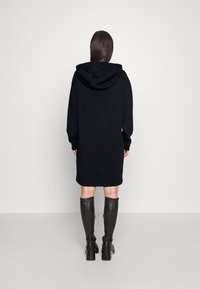 Schwarzes Hoodie-Kleid mit Kapuze, langen Ärmeln und geripptem Saum. Kombiniert mit kniehohen schwarzen Stiefeletten mit Absatz. Einfarbiger Hintergrund.