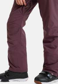 Fialové lyžařské kalhoty s elegantním designem, vybavené zipovou kapsou a zesíleným švem; v kombinaci s černými sněhulkami a bílou podrážkou.