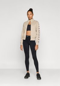 Beige glidelåsjakke med høy krage, svart sportstopp, svarte leggings og svarte sko. Jakken har logo og mønstrede detaljer på ermene.