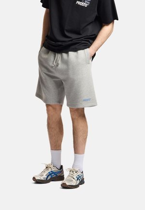 Osoba nosząca jasnoszorty z troczkami i logo "PROSTO", białe skarpetki za kostkę, beżowo-niebieskie buty do biegania oraz czarną koszulkę.