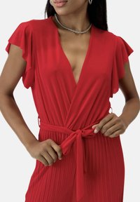 Vestido rojo con mangas cortas de volantes, escote en V y cintura ajustada con lazo. Textura plisada que se extiende a lo largo. Tela suave y ligera.