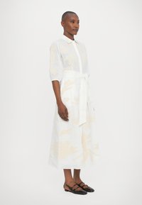 White linen dress with light beige floral pattern, short puff sleeves, a collar, and a tied belt at the waist. Worn with black flats.