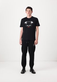 Black t-shirt with a white graphic of two dog heads and "HUGO" in red. Paired with black sweatpants and black shoes, standing against a white backdrop.
