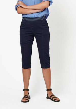 Marineblaue Cropped-Hosen aus weichem Stoff, mit flachem Bund und genähten Details. Kombiniert mit schwarzen flachen Sandalen.