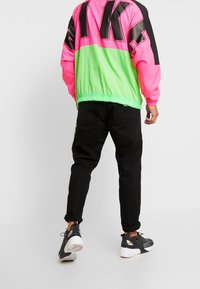 Man som bär en neonrosa och grön jacka, svarta uppvikta byxor och svarta och vita sneakers som går framför en helt vit bakgrund.