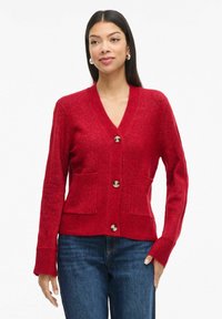 Žena nosi crveni kardigan na dugmad s dvije prednje džepove i plave traperice, stojeći nasuprot jednostavne bijele pozadine.