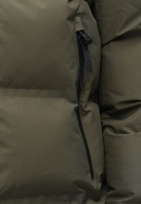 Olívazöld steppelt kabát részlet, quiltelt textúrával, sima anyaggal és fekete cipzáras zsebbel, amely márkajelzéssel van ellátva.