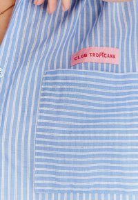Jasnoniebieska koszula w paski z miękkiego materiału, z kieszonką na lewej piersi i różową metką z napisem "CLUB TROPICANA." Widoczne poziome paski.