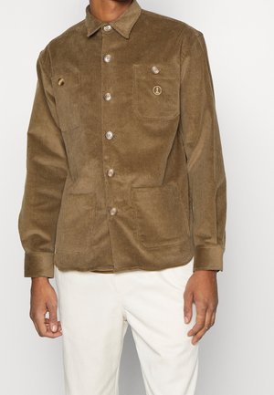 Veste en velours marron avec de gros boutons et trois poches devant, portée avec un pantalon blanc sur une personne devant un fond uni.