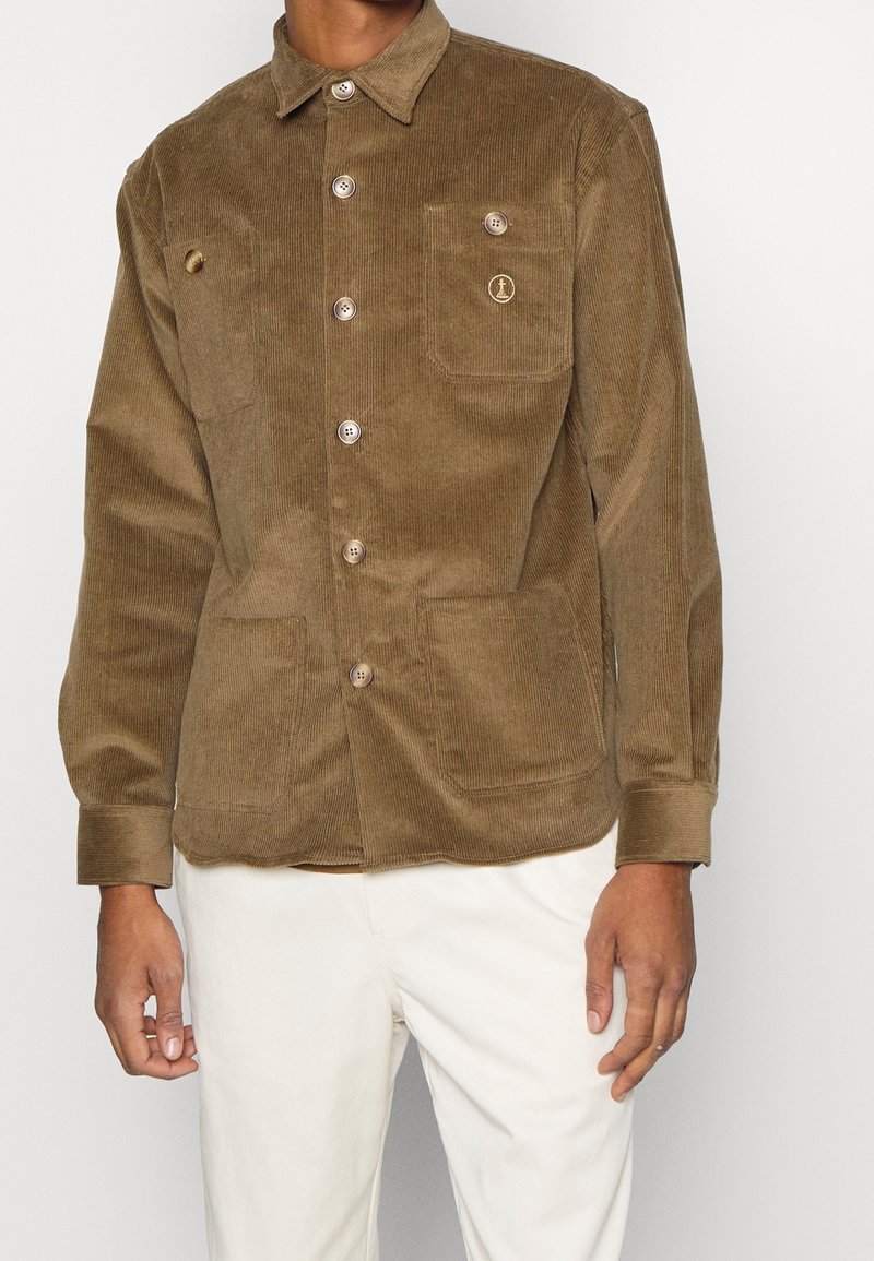 Veste en velours marron avec de gros boutons et trois poches devant, portée avec un pantalon blanc sur une personne devant un fond uni.