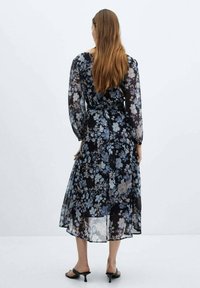 Vestido maxi negro con estampado floral, mangas largas y transparentes, y cintura con lazo. Presenta una mezcla de flores azules y crema, tejido semitransparente y una silueta fluida.