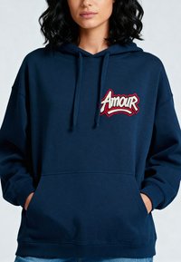 Felpa blu navy con cappuccio regolabile e tasca frontale. Ricamo "Amour" in bianco e rosso. Materiale morbido e texturizzato.