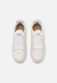 Zapatillas blancas con parte superior de cuero suave, detalles perforados, lengüeta acolchada y plantilla marrón. Cuenta con cordones blancos y acentos con el logotipo.