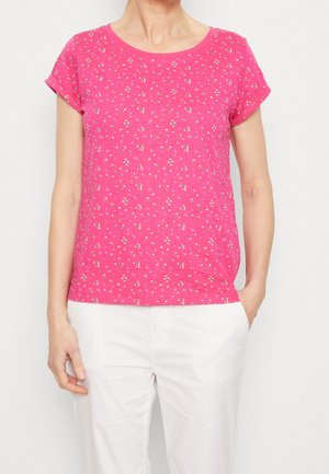 Kobieta ubrana w różową koszulkę z krótkim rękawem, ozdobioną małymi białymi kwiatowymi wzorami, oraz białe spodnie, stojąca z jedną ręką w kieszeni.