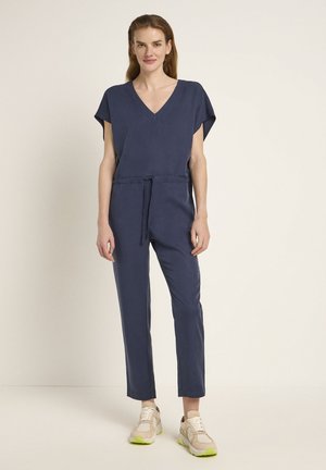 Marineblauer Jumpsuit mit kurzen Ärmeln, V-Ausschnitt und Kordelzug in der Taille. Glatter Stoff und schmal zulaufende Beine. Kombiniert mit leichten Sneakern mit neonfarbenen Akzenten.