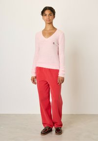 Un pull léger en rose clair, fluffy, avec un col en V et une petite patch colorée, associé à un pantalon en velours côtelé rouge vif et des chaussures brunes à lacets.