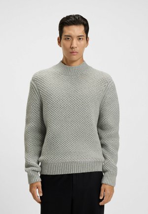 Uomo che indossa un maglione lavorato a maglia grigio chiaro con colletto alto a coste e pantaloni neri, in piedi contro uno sfondo bianco semplice.