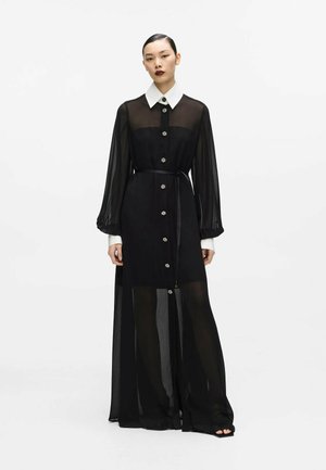 Femme portant une longue robe noire transparente avec un col blanc et des poignets blancs, boutonnée devant, avec une ceinture à la taille, debout sur un fond uni.