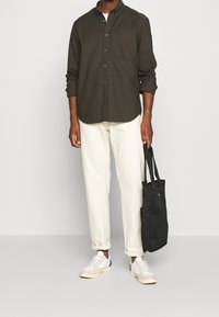 Camicia button-up verde scuro con tasca sul petto, abbinata a pantaloni bianchi e sneakers bianchi. Una borsa tote nera è tenuta nella mano destra.