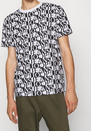 Mann trägt ein schwarz-weißes kurzärmeliges Hemd mit durchgehendem "JOOP! JEANS" Schriftzugmuster und olivgrüne Hose vor einfachem Hintergrund.