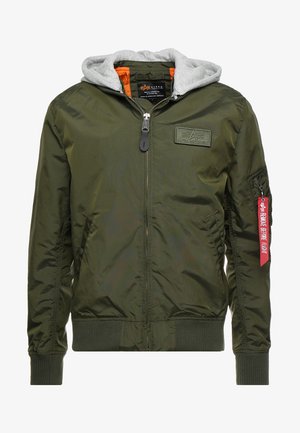 Olijfgroene bomberjack met een grijze capuchon, een voorrits, ribgebreide manchetten en een badge op de borst. Voorzien van een rode tag met de tekst "VERWIJDER VOORDAT JE VLUCHT."