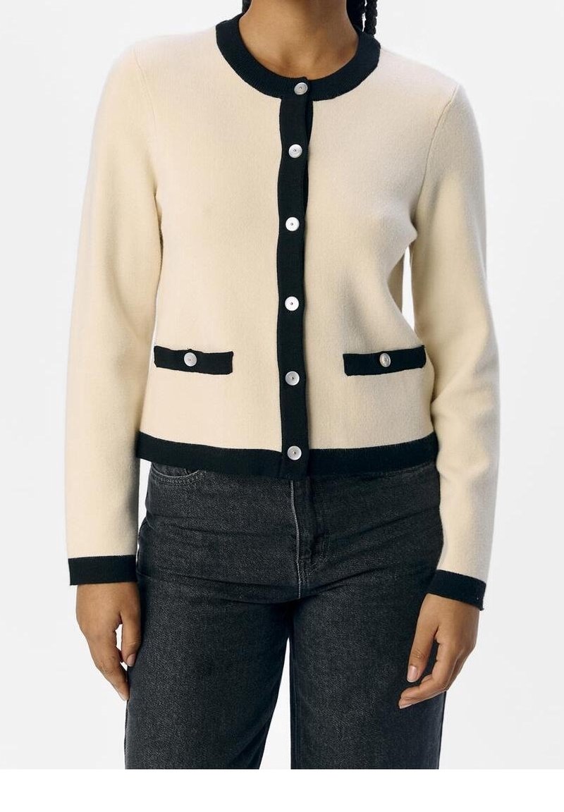Beige stickad cardigan med svart kant, med rund halsringning, knäppning fram och två främre fickor med dekorativa svarta detaljer.