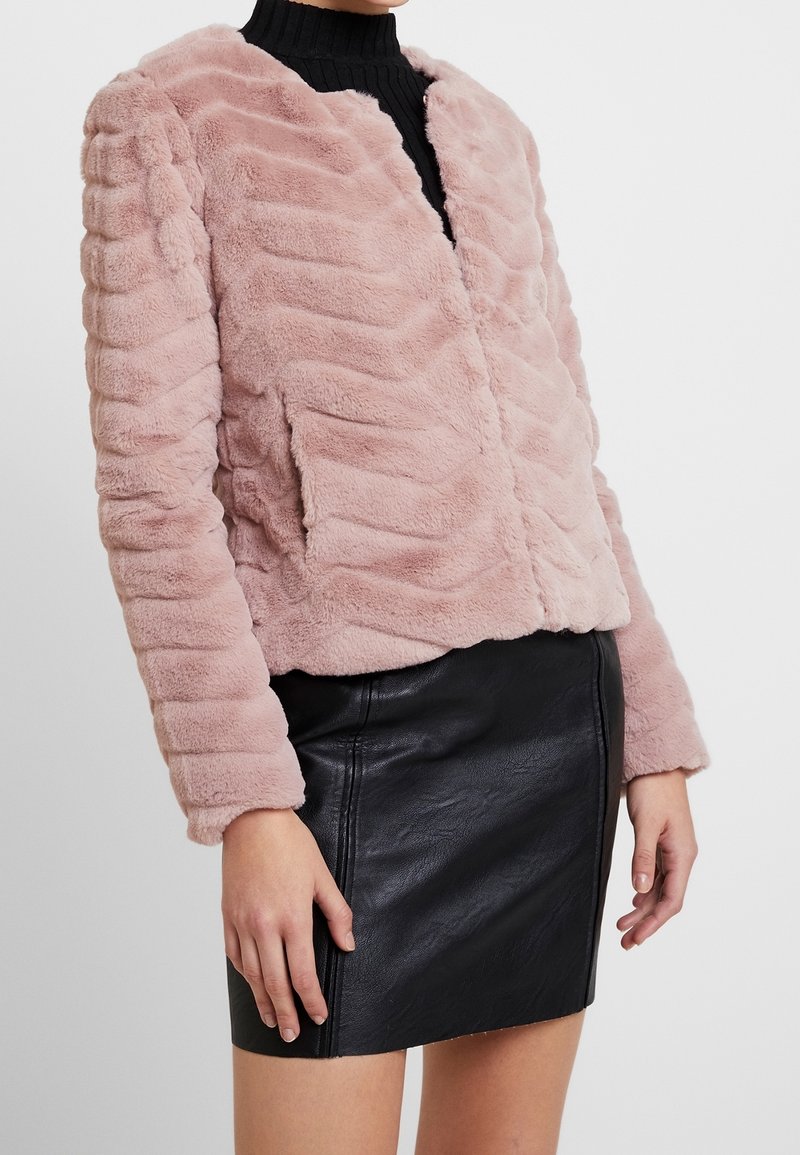 Sztuczny futrzany płaszcz w miękkim różowym kolorze z poziomą fakturą prążkowaną, z zapięciem na suwak z przodu i długimi rękawami, w połączeniu z czarną skórzaną mini spódnicą.