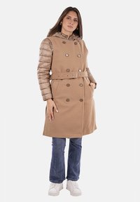 Cappotto beige doppiopetto con maniche imbottite, cintura in vita, tasche laterali e cappuccio. Indossato sopra jeans blu e sneakers bianche.