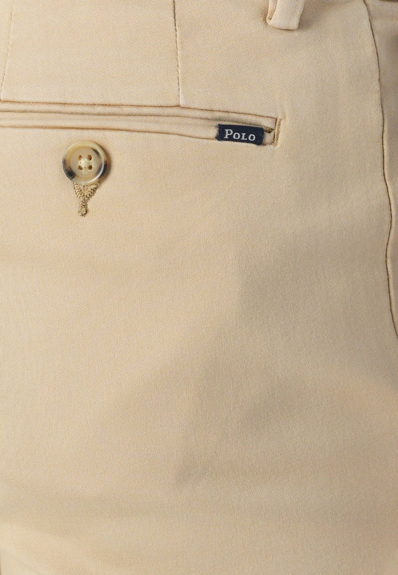 Beżowe spodnie o gładkiej fakturze materiału, z guzikiem i małą metką "Polo". Design obejmuje przednią kieszeń z dodatkowymi detalami szewnymi.