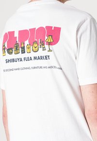 Hvid bomulds T-shirt med et farverigt grafisk design på ryggen, herunder ikoner såsom flasker, en lampe og et ur, med tekst nedenunder.
