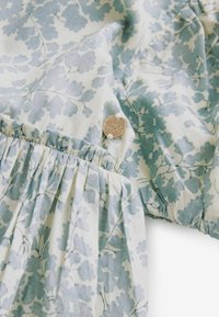 Cremfarbener Stoff mit blauem Blumenmuster, ausgestattet mit geraffter Textur und einem Metallknopfakzent. Weiches, leichtes Material.