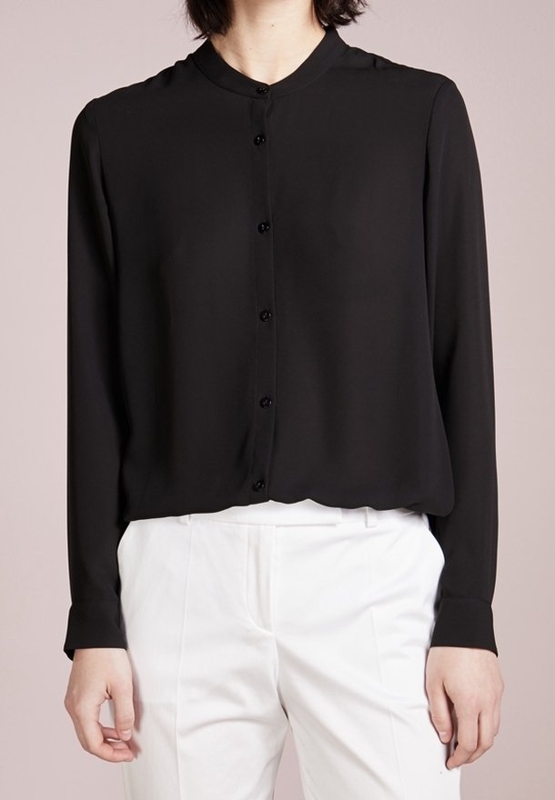 Personne portant une chemise noire à manches longues boutonnée avec un pantalon blanc, debout devant un fond clair uni.