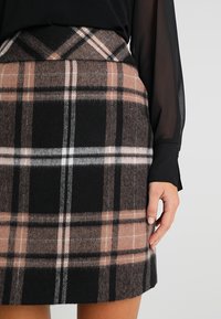 Femme portant une jupe à carreaux marron, noire et blanche mi-longue, avec un chemisier noir transparent à manches longues, la main posée sur le côté de la jupe.