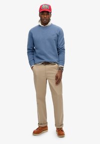 Lichtblauwe sweatshirt over een grijze overhemd met kraag, khaki broek, bruine leren schoenen en een rode pet met logo; casual fit, gestructureerde stof.