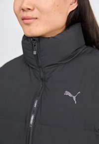 Doudoune noire avec un col haut, fermeture éclair et un logo Puma réfléchissant. Texture lisse, design matelassé et tissu léger.