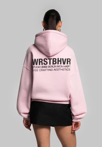 Felpa con cappuccio rosa dal fit rilassato, con testo nero sul retro che recita "WRSTBHVR" e ulteriori dettagli di design sottostanti.