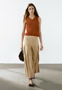 Top senza maniche a scollo a V ruggine abbinato a pantaloni a palazzo beige con pieghe; tiene in mano una borsa marrone testurizzata; indossa sandali neri piatti.