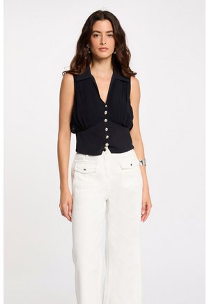 Femme aux longs cheveux noirs portant un chemisier noir sans manches à boutons et un pantalon blanc taille haute avec poches avant, debout devant un fond uni.