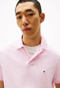 Lichtroze poloshirt van getextureerde stof, met een kraag en een klein geborduurd logo op de borst.