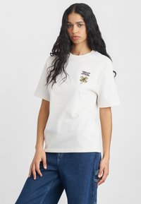 Biela bavlnená tričko s krátkymi ruk�ávmi, s vyšitým zeleným a žltým dizajnom štvorlístka a textom na hrudi.