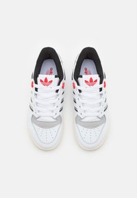 White athletic shoes with black stripes and grey accents. Features round red eyelets and textured black padding around the collar.