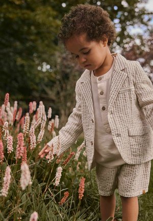 Enfant aux cheveux bouclés portant un costume à motifs touchant délicatement des fleurs roses et blanches dans un jardin avec un arrière-plan de verdure floue.