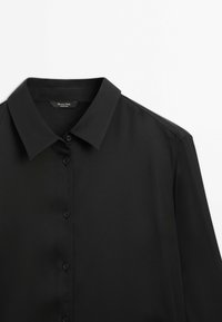 Chemise noire à manches longues avec boutons et col pointu, en tissu lisse, présentée sur un fond blanc.
