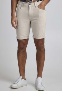 Beige denimshorts med rullkanter, med frontlommer og en subtil strukturert overflate, paret med hvite sneakers.