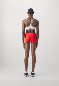 Sportos öltözet, amely tartalmaz egy fehér sportmelltartót fekete díszítéssel, piros sortot és fekete sportcipőt. A hátsó nézet jól látható, hogy testhezálló tervezésű és pántos.
