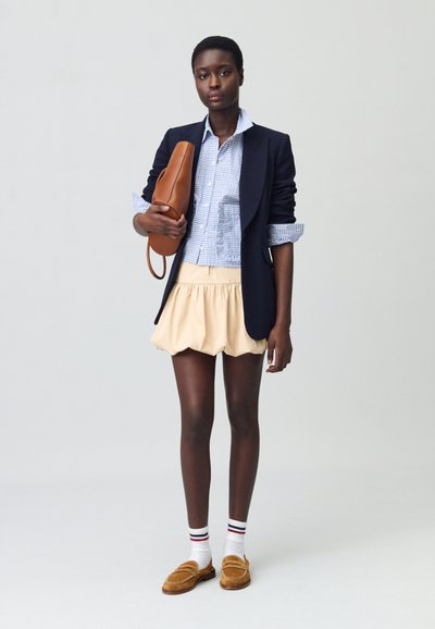 Blazer bleu marine sur une chemise à carreaux bleus, jupe crème, sac marron et mocassins beiges ; chaussettes blanches avec des rayures rouges et bleues marines.