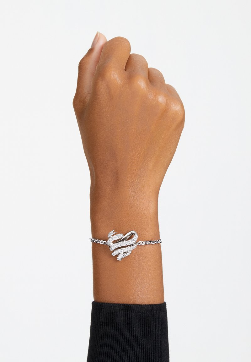 Strieborný náramok s dizajnom v tvare srdca, ktorý má zložitú detalizáciu a textúrovaný reťazec. Nosený na ruke s neutrálnym odtieňom.
